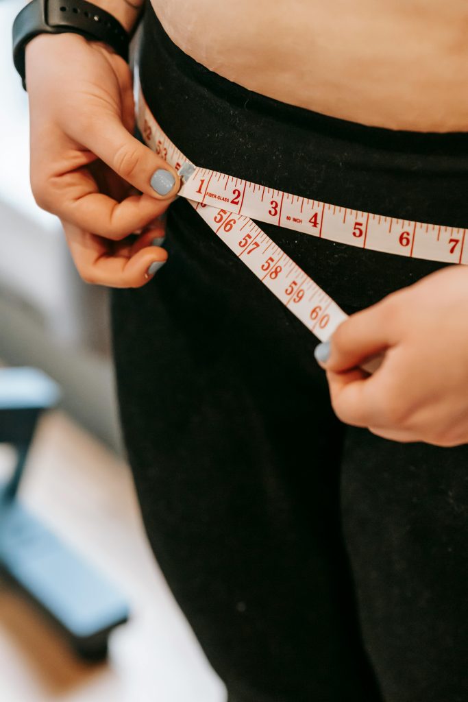 Home From above of crop anonymous plump female using measuring tape around hips in gym